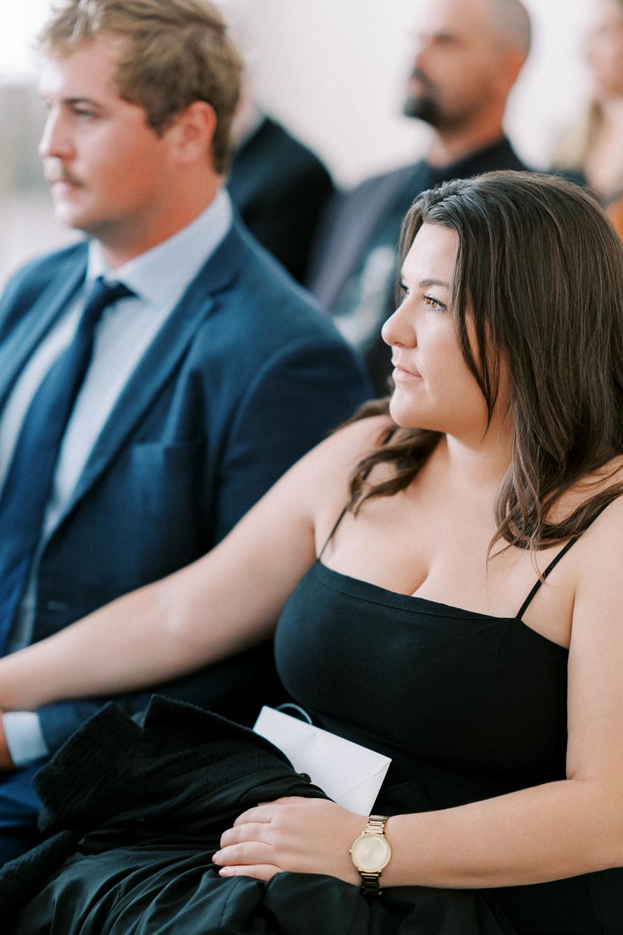 A woman in a black dress sits attentively at a formal event, with a man in a blue suit beside her, both engaged in watching the proceedings.