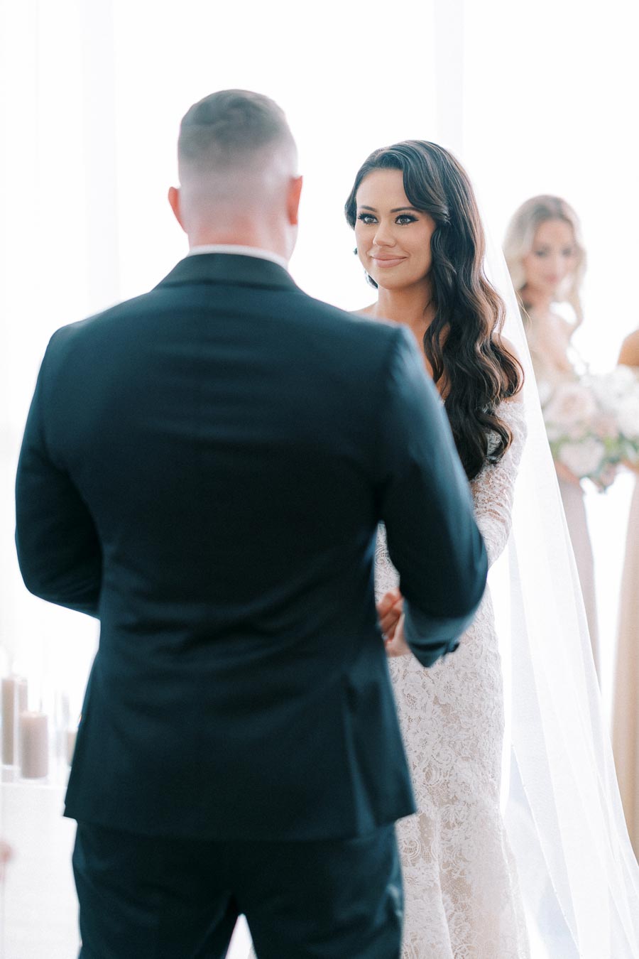 A bride in a lace dress and veil lovingly gazes at her groom during their wedding ceremony, with a bridesmaid holding a bouquet in the background.