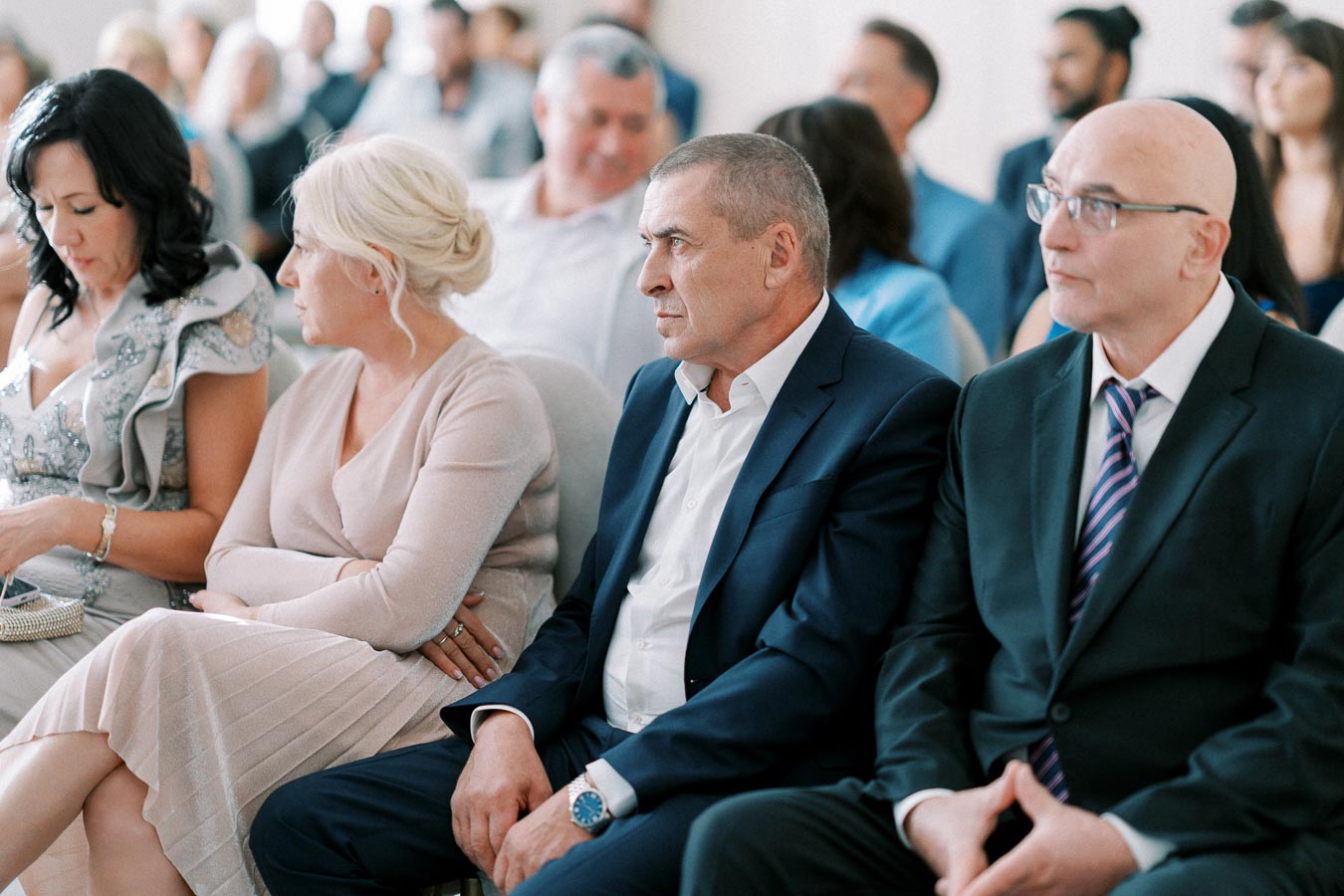 A group of people seated in an event, wearing formal attire, attentively listening. The image highlights a diverse audience in a professional setting, conveying a sense of engagement and focus.
