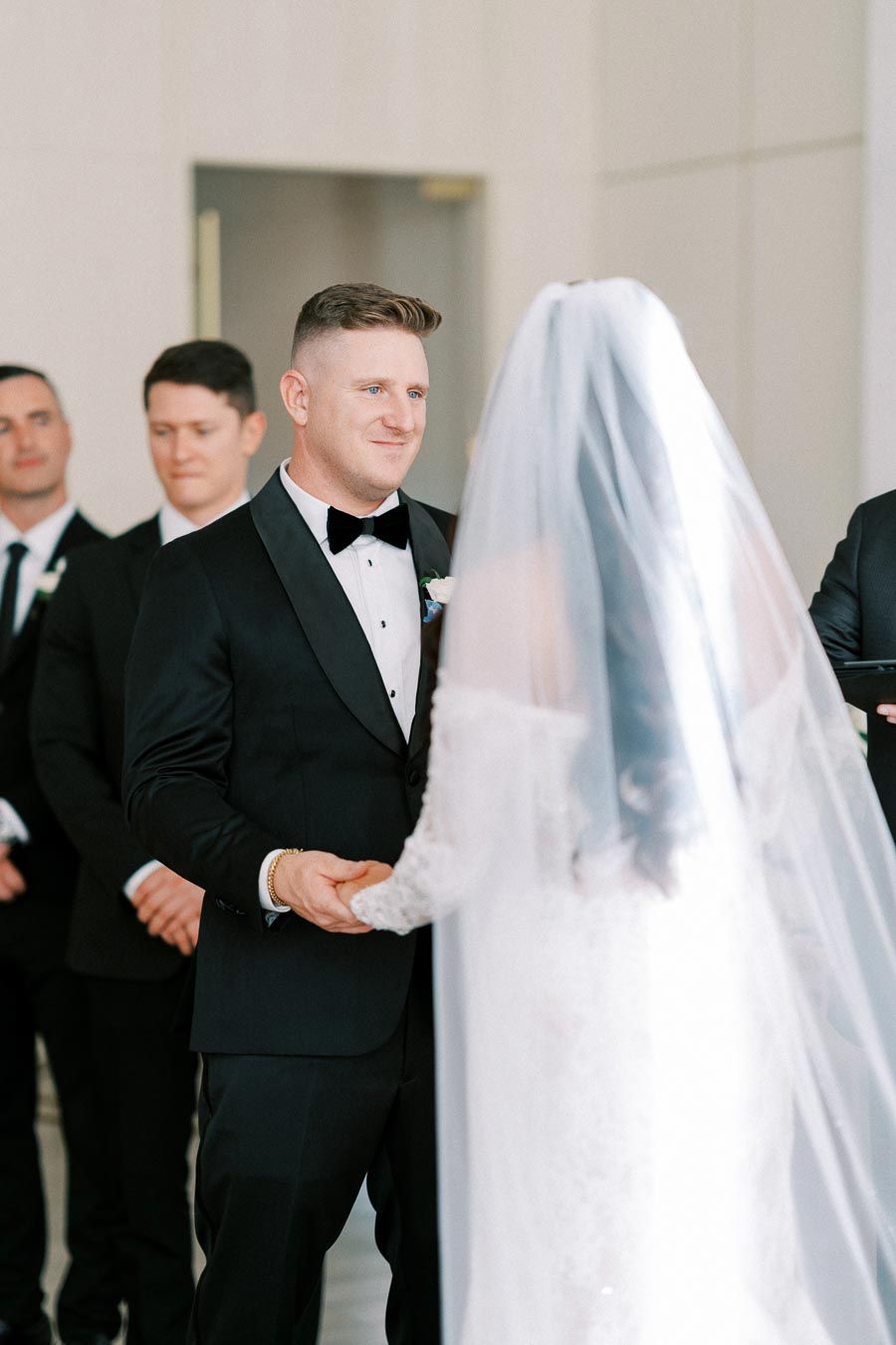 Wedding ceremony with groom in black tuxedo smiling at bride in white gown and veil, surrounded by groomsmen in a bright event space.