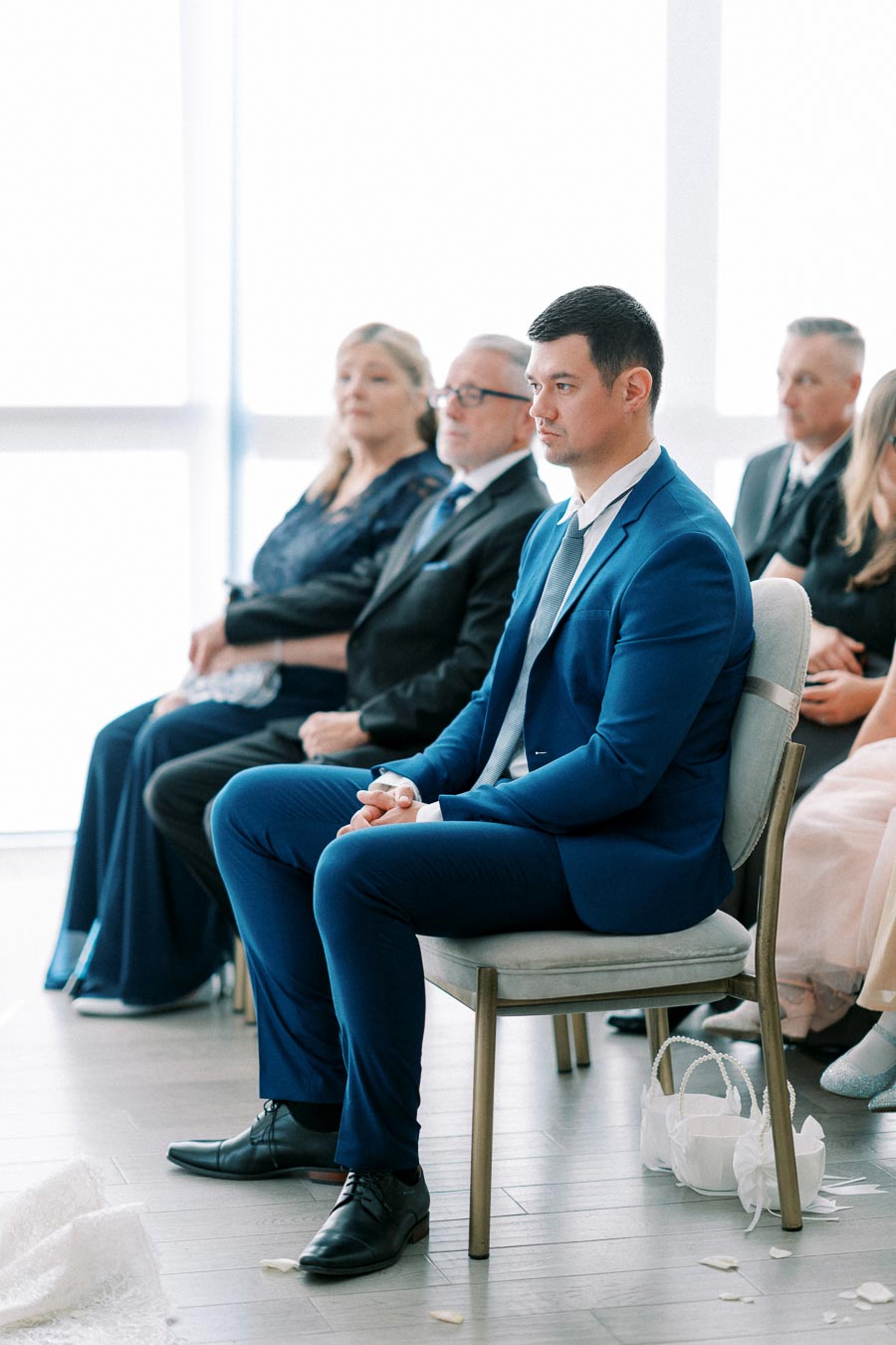 A group of formally dressed individuals seated at an indoor event; a man in a blue suit sits attentively in the foreground, with others behind him, suggesting a wedding or formal ceremony setting.