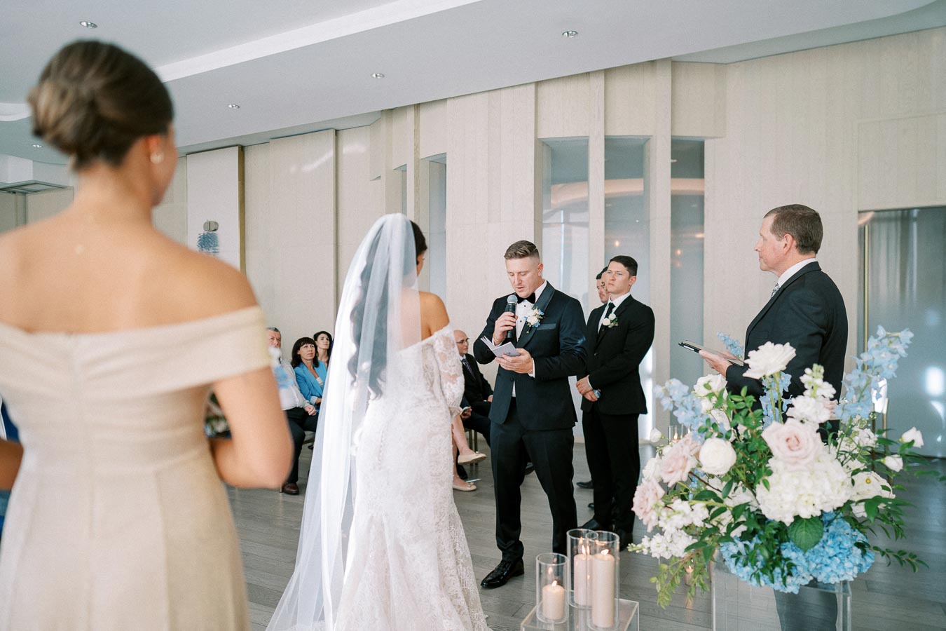 Wedding ceremony with groom reading vows, bride in lace gown, and officiant. Elegant indoor setting decorated with flowers and candles.