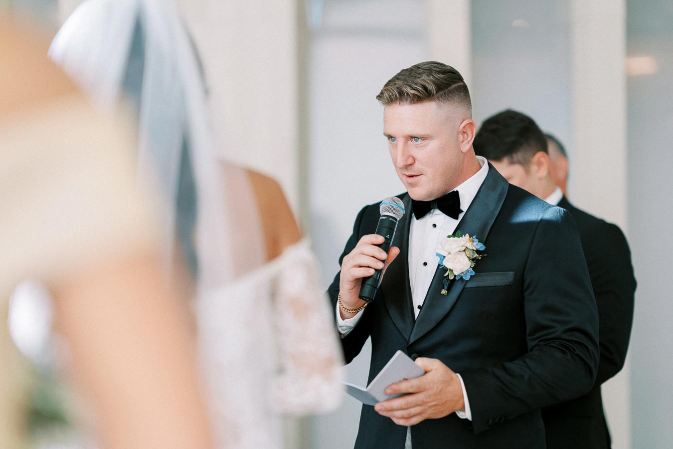 A groom in a black tuxedo holds a microphone and vows booklet while delivering a speech during a wedding ceremony.