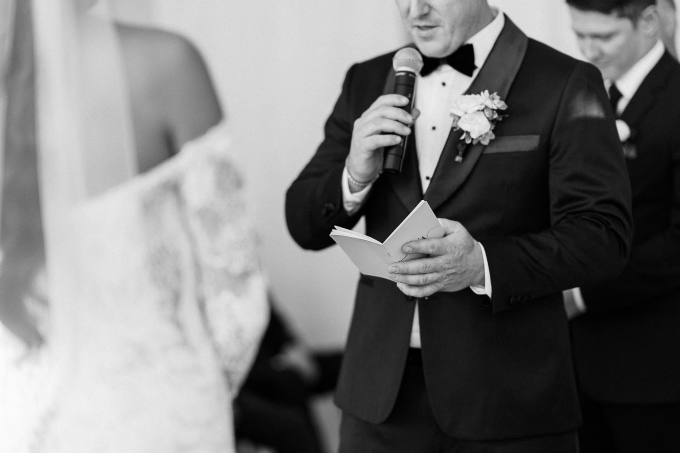 A groom in a tuxedo reads vows from a notebook while holding a microphone during a wedding ceremony.