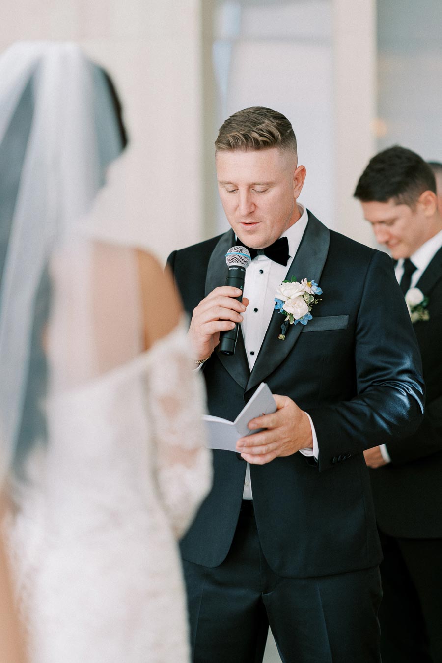 Groom in a black tuxedo holding a microphone, reading vows during a wedding ceremony, with the bride in a white dress standing opposite him.