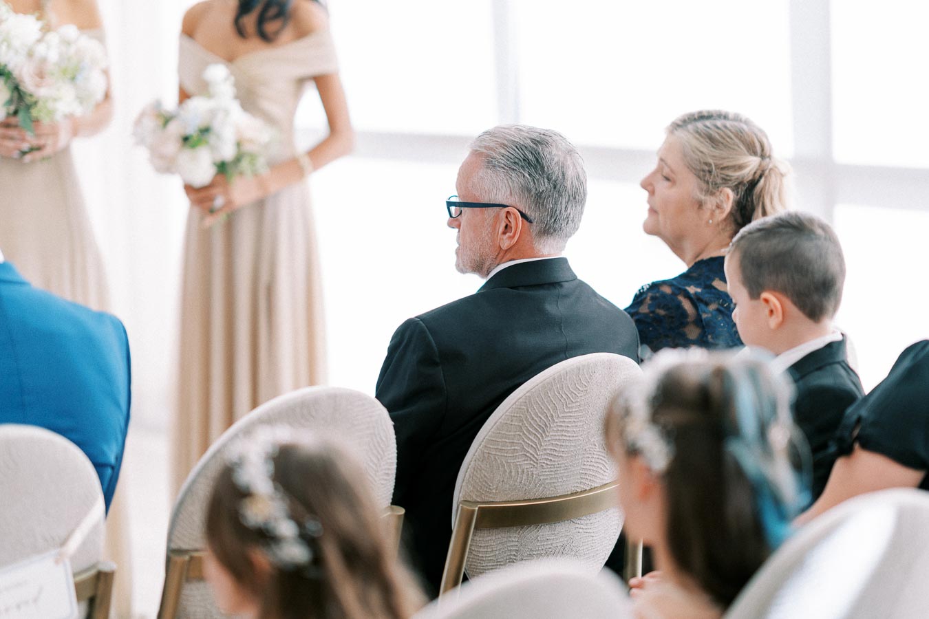 A small group of elegantly dressed wedding guests sitting and attentively watching a ceremony, with a child in the foreground and bridesmaids visible in the background holding bouquets.