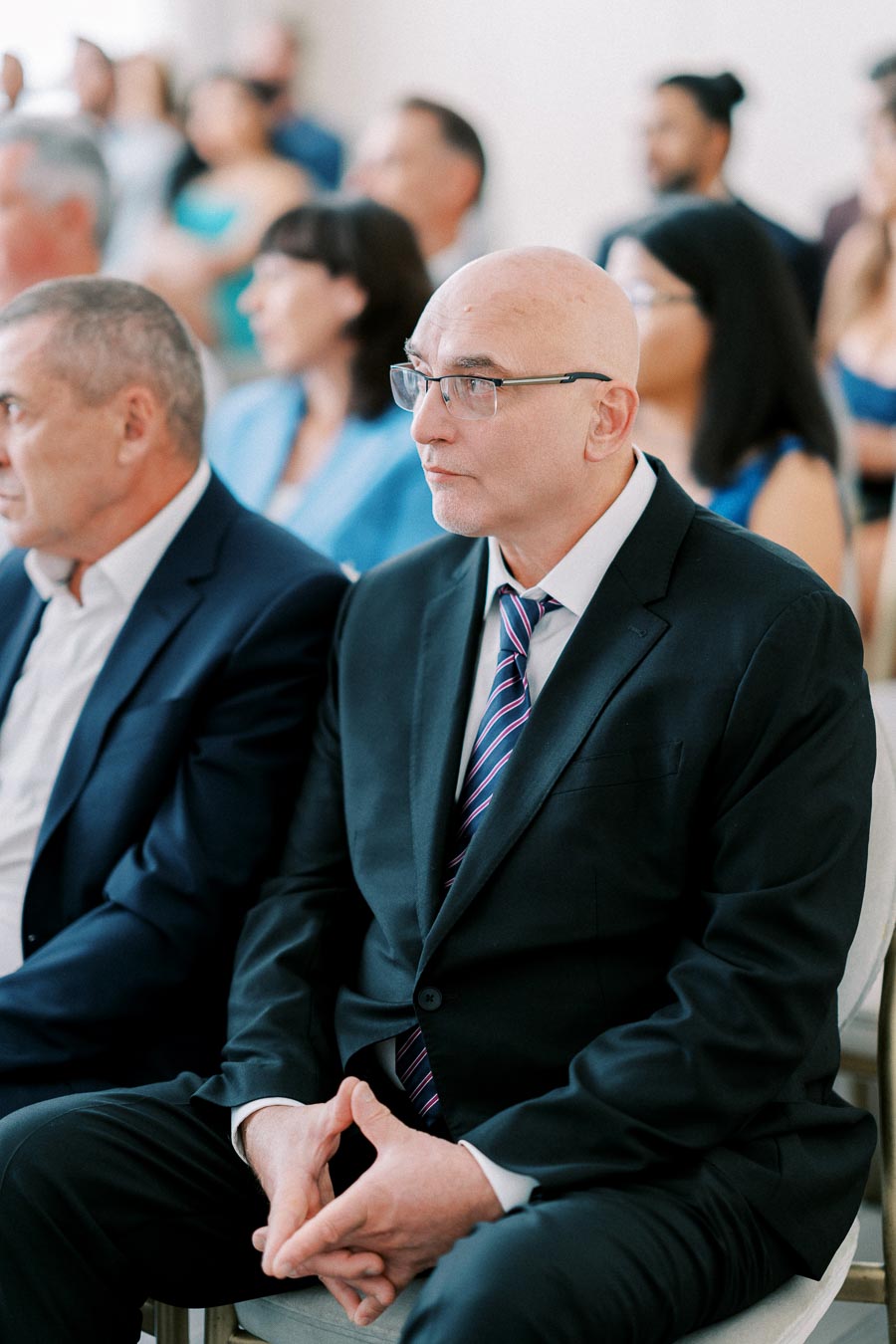 A group of people attending a formal event, with a man in a black suit and tie sitting attentively in the foreground.