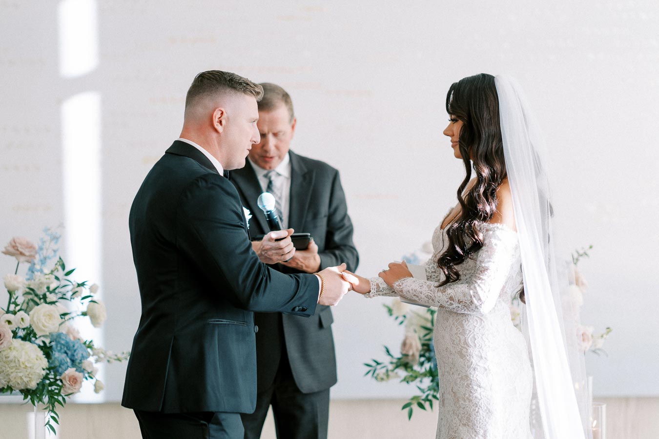 A bride and groom exchanging vows during a wedding ceremony, with a minister officiating. The bride wears a lace wedding dress with a veil, and the groom is in a black suit, surrounded by floral arrangements.