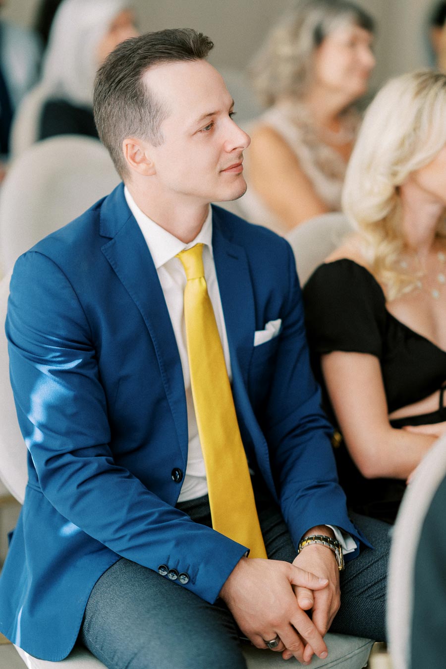 A man in a blue suit with a yellow tie attentively listens during an event, seated among other attendees.