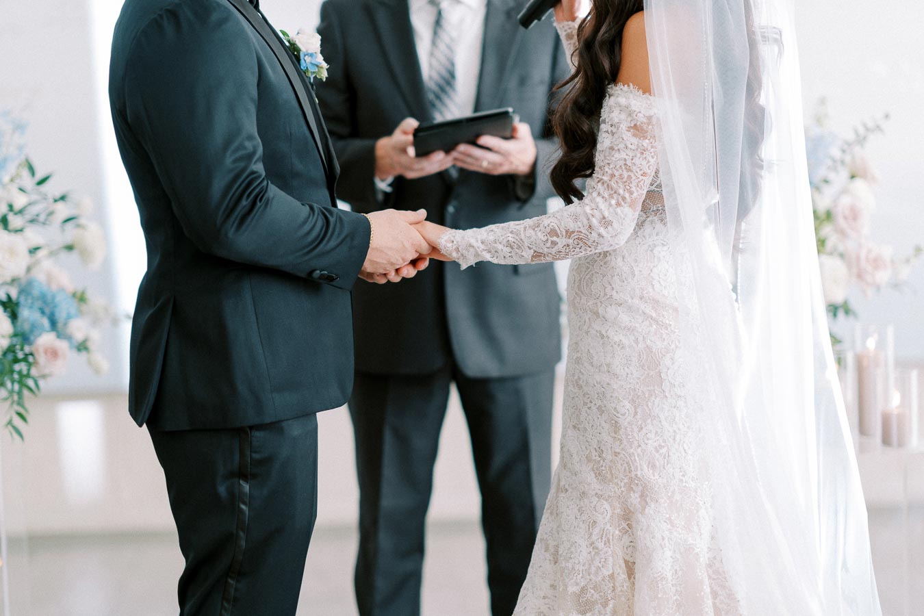 Bride and groom holding hands during wedding ceremony with officiant in the background.