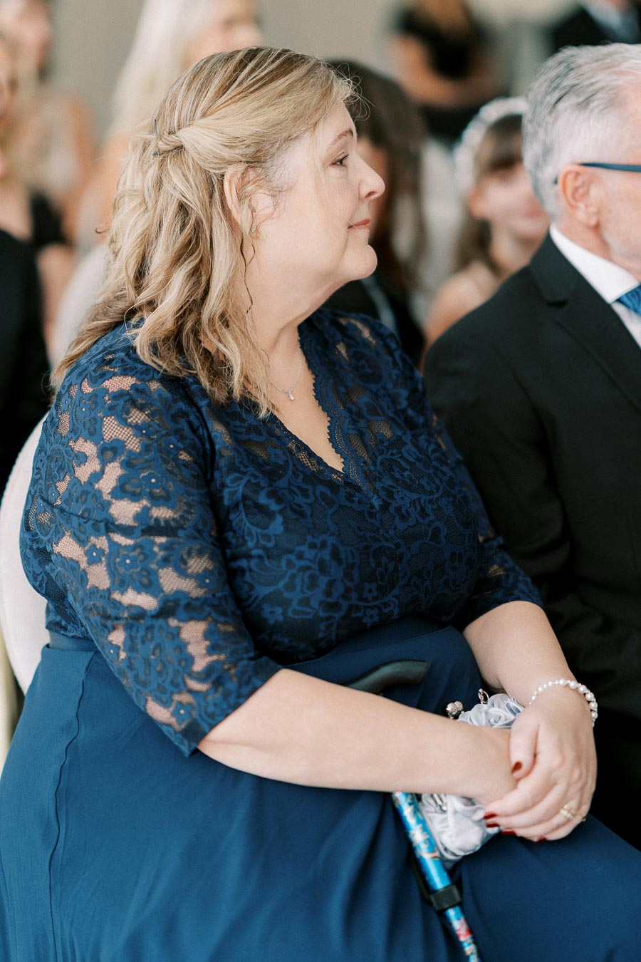 A woman in a navy lace dress sits attentively at a formal event, holding a small bouquet and wearing pearl jewelry.