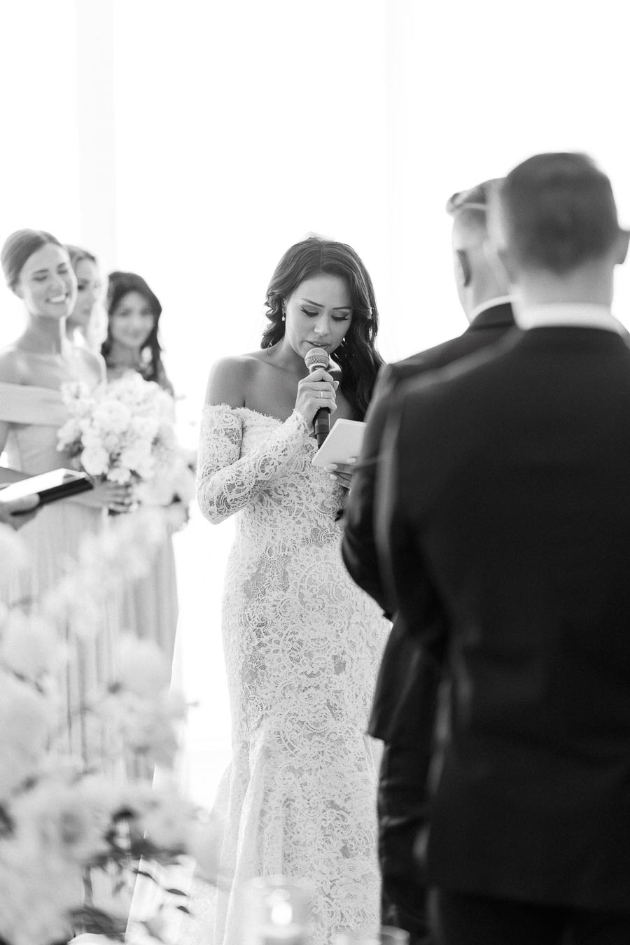 A bride in a lace wedding dress delivers a heartfelt speech during a wedding ceremony, surrounded by bridesmaids holding bouquets.