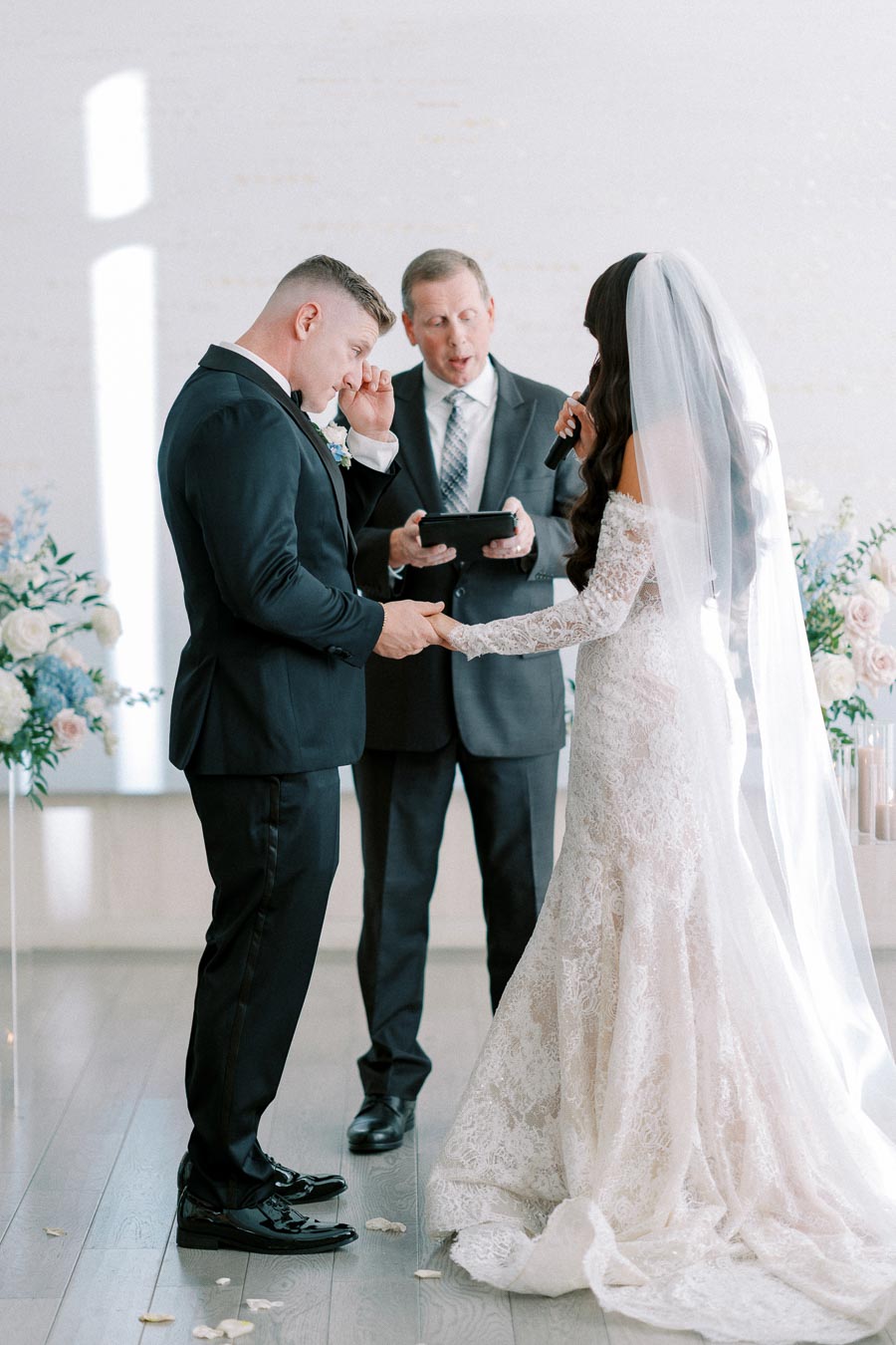 A bride and groom stand facing each other during an indoor wedding ceremony, with an officiant holding a book and floral decorations in the background. The bride wears a lace gown with a long veil, while the groom is dressed in a black suit.