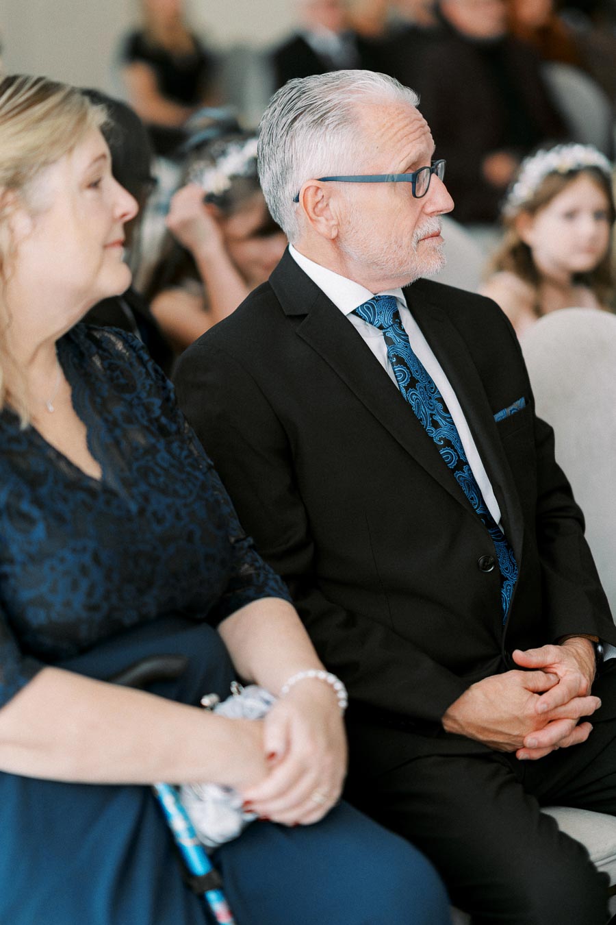 An elderly couple dressed in formal attire, seated at a wedding ceremony, looking attentively forward; the man wearing a suit with a patterned blue tie and glasses, and the woman in a navy lace dress holding a walking cane.