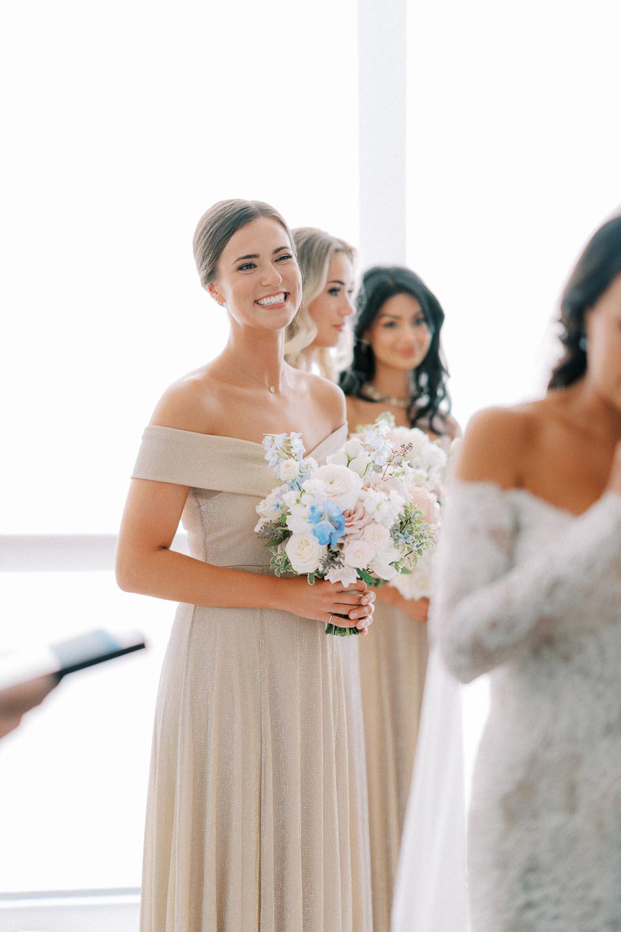 A smiling bridesmaid in an elegant off-shoulder dress holding a bouquet of pastel flowers at a bright indoor wedding ceremony.