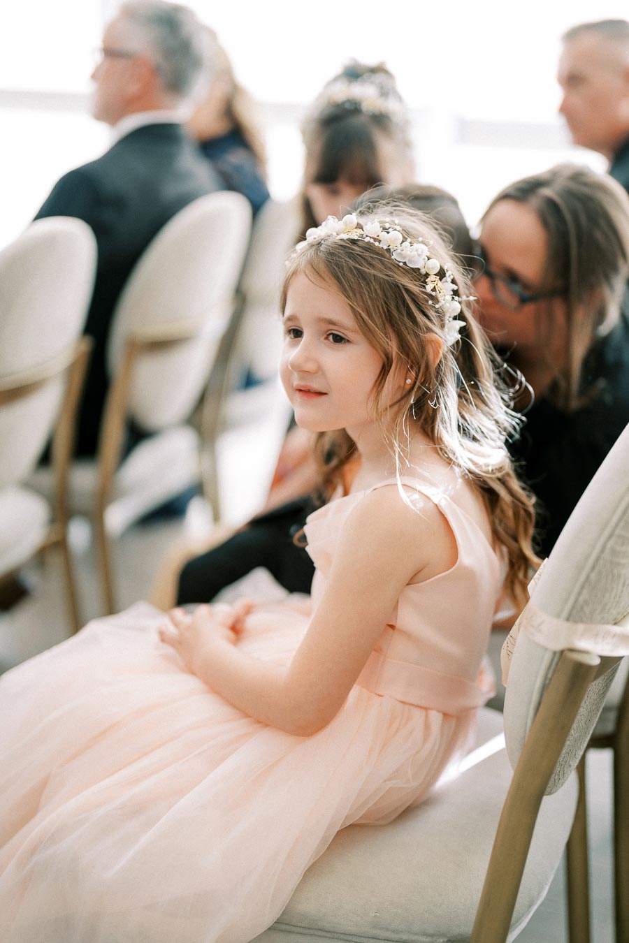 Alt text: Young girl in a pastel pink dress with a floral headband seated at a formal event, other attendees visible in the background.