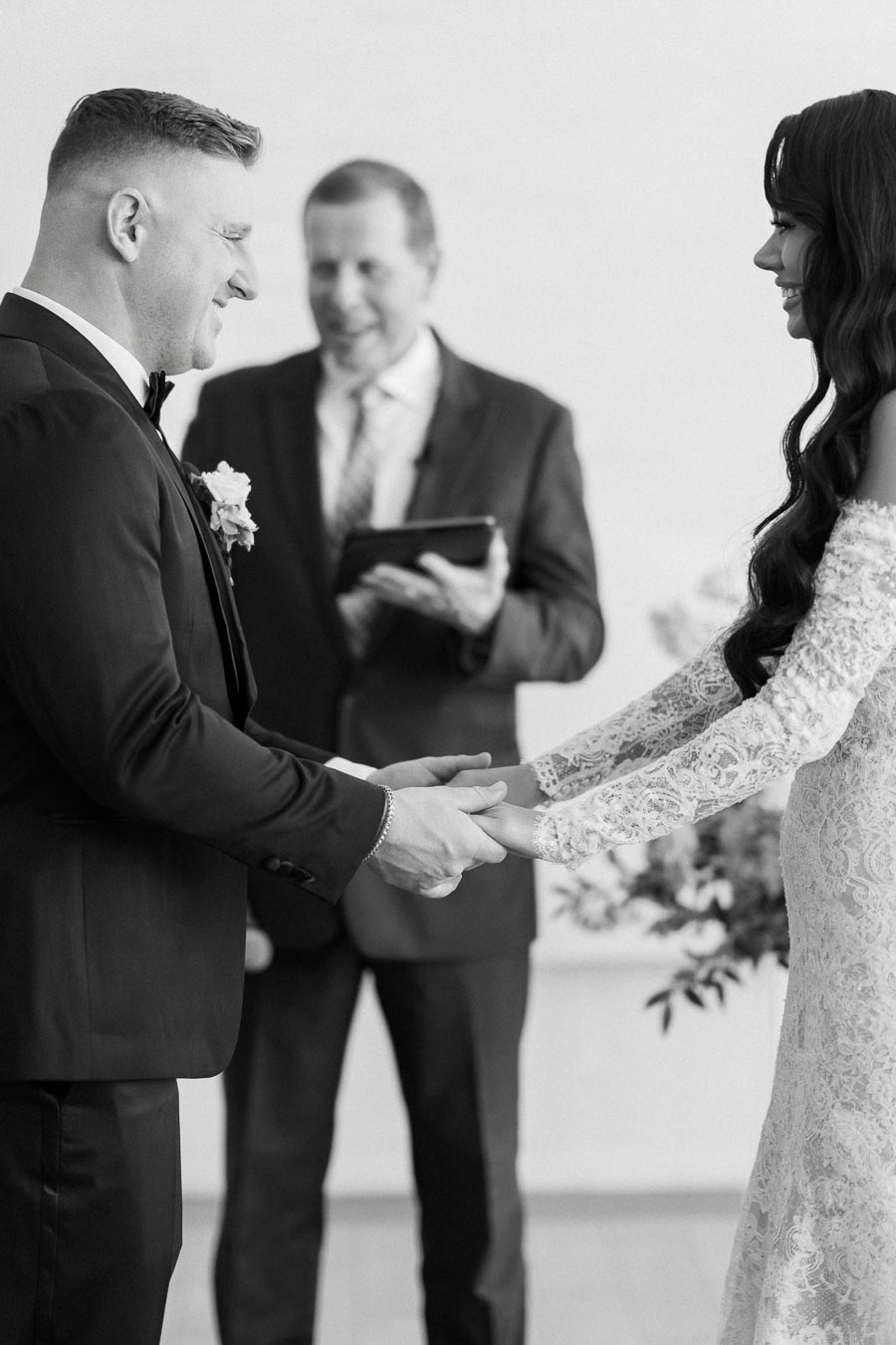 Black and white image of a couple holding hands during their wedding ceremony, with an officiant in the background, in an elegant indoor setting.