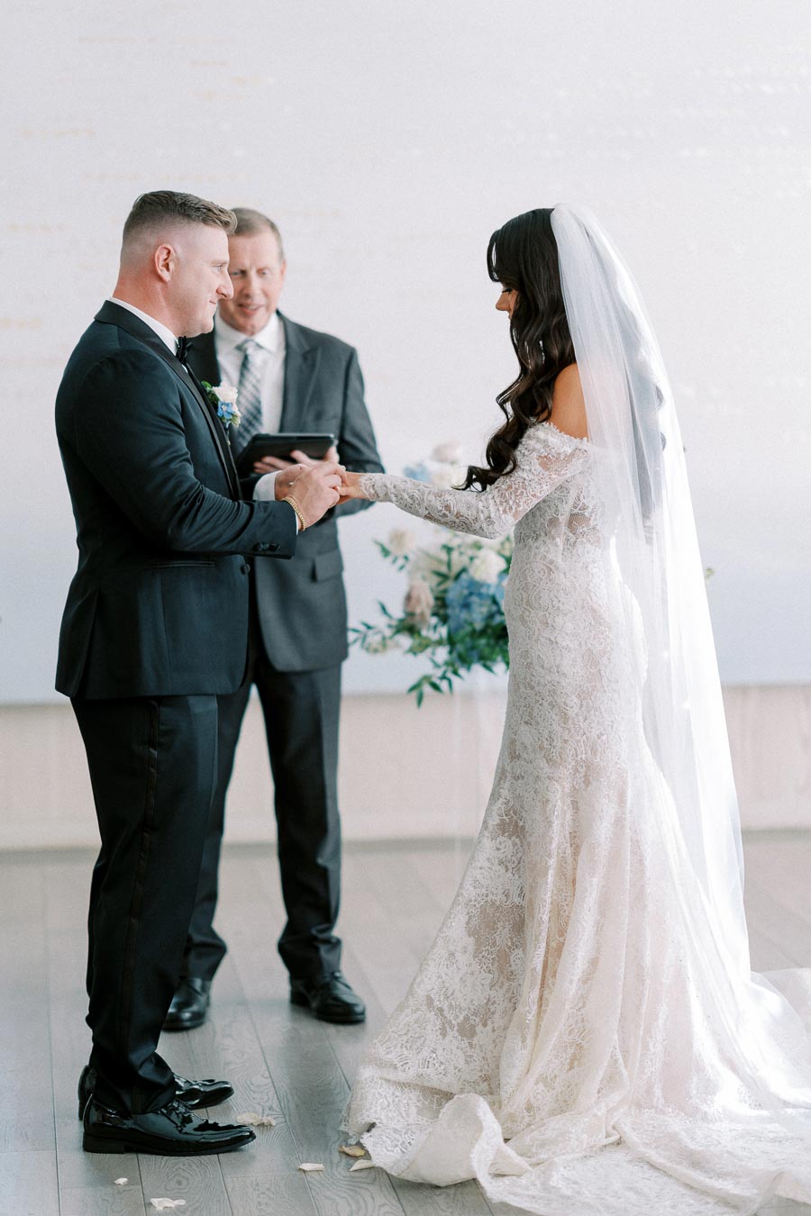 Bride and groom exchanging vows during wedding ceremony, with officiant present; bride in lace gown and veil, groom in black tuxedo.