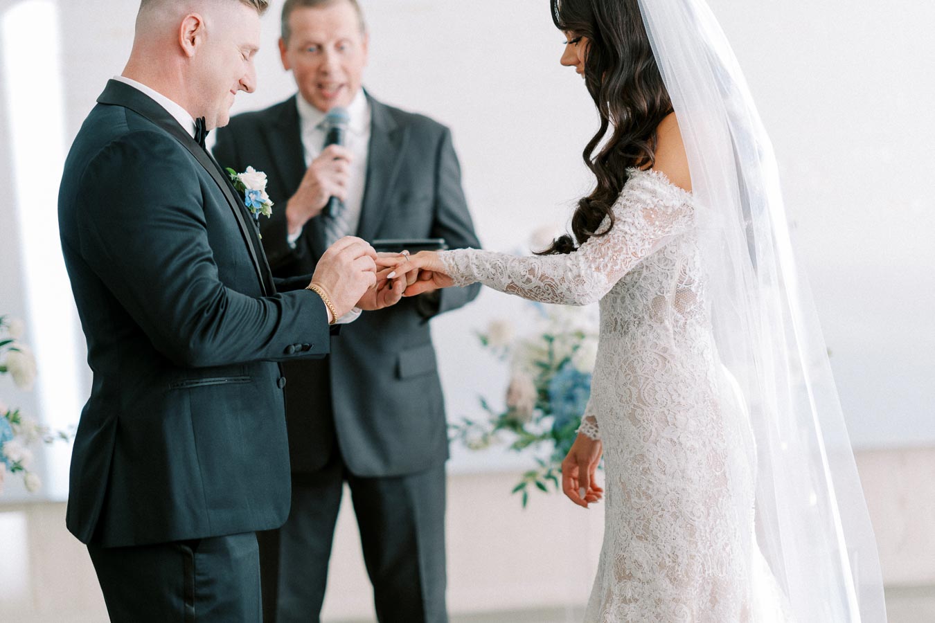 A groom places a wedding ring on the bride's finger during a ceremony, with an officiant in the background. The bride is wearing a lace wedding dress and veil, while the groom is dressed in a formal suit.