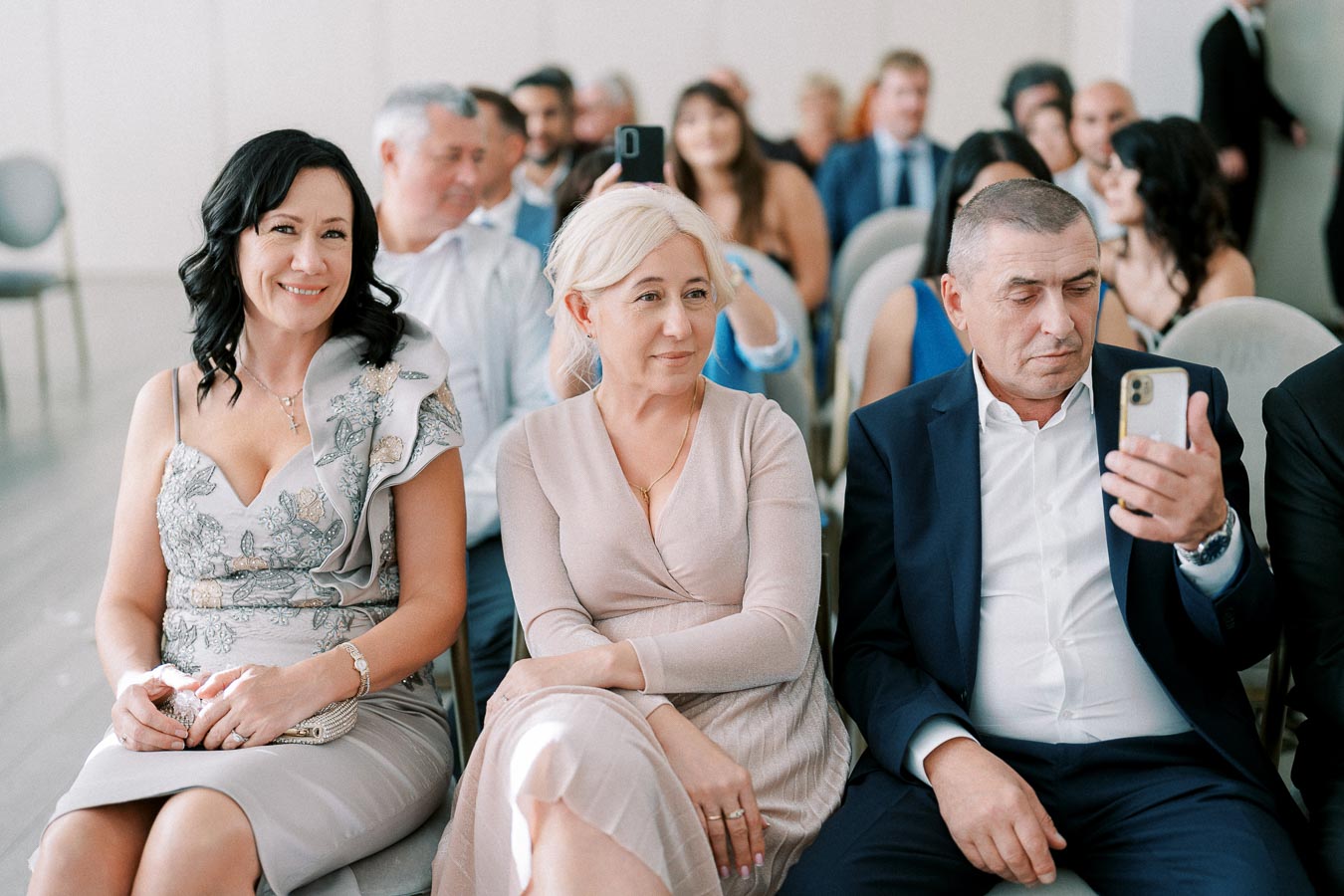 A group of elegantly dressed people seated at an indoor event, with a man using a smartphone and others smiling and engaged in conversation, creating a warm and social atmosphere.