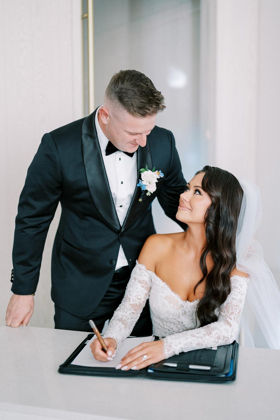 Bride in elegant lace wedding dress signing marriage documents at ceremony table while groom in black tuxedo gazes lovingly, symbolizing matrimonial commitment and joy.