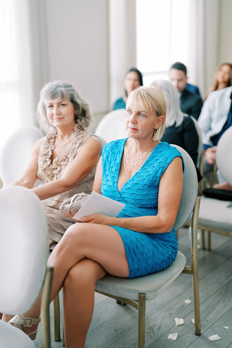 Two women seated in a bright, elegant setting, attentively listening to an event. The woman in the foreground is wearing a vibrant blue dress, holding a program, while the other woman wears a beige dress. The background shows other attendees slightly blurred, suggesting focus on the main subjects.