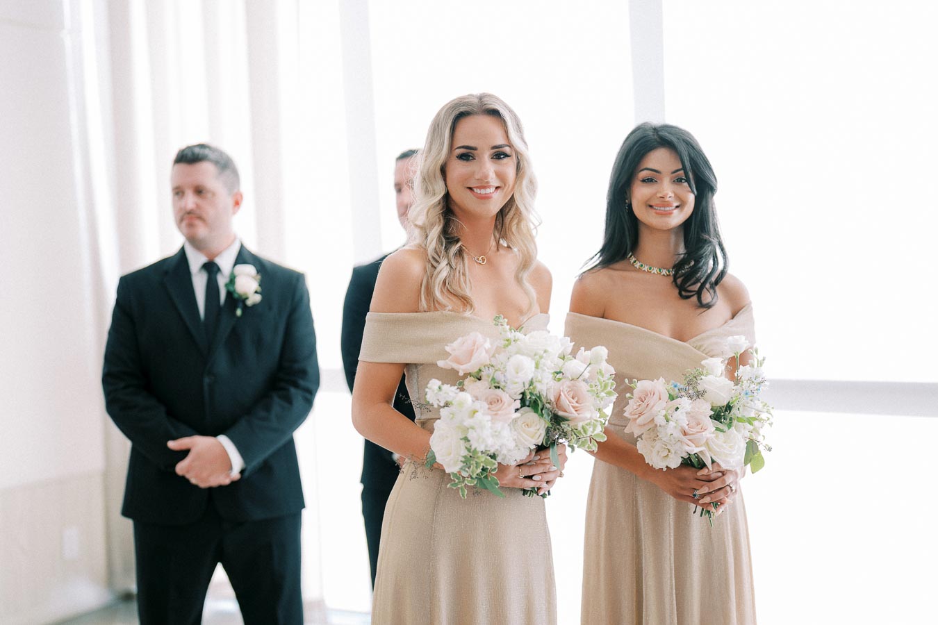 Bridesmaids in elegant off-the-shoulder dresses holding pastel floral bouquets standing with groomsmen in a bright wedding venue.