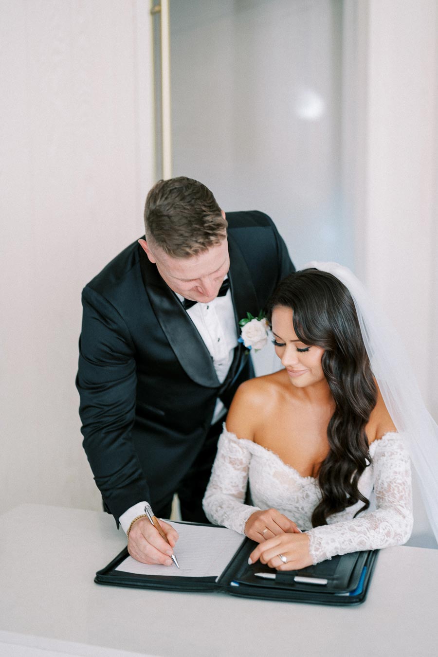 A groom in a black tuxedo signing a document while the bride in a white lace gown and veil looks on, symbolizing a joyous wedding moment.