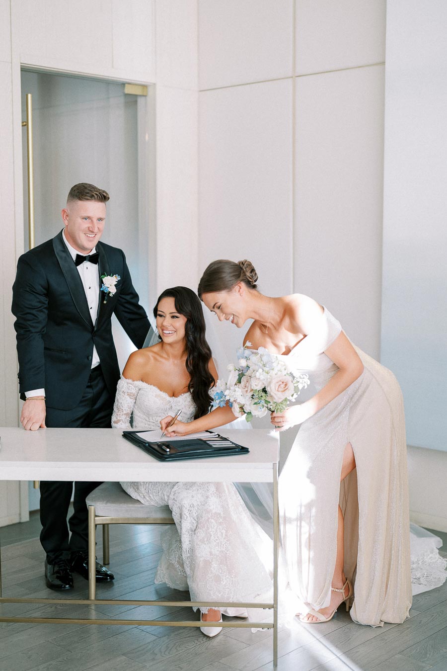 Three people in formal attire during a wedding ceremony. A smiling bride sits while another woman signs a document, and a man in a tuxedo stands beside them. The room is elegantly decorated, with natural light enhancing the scene.