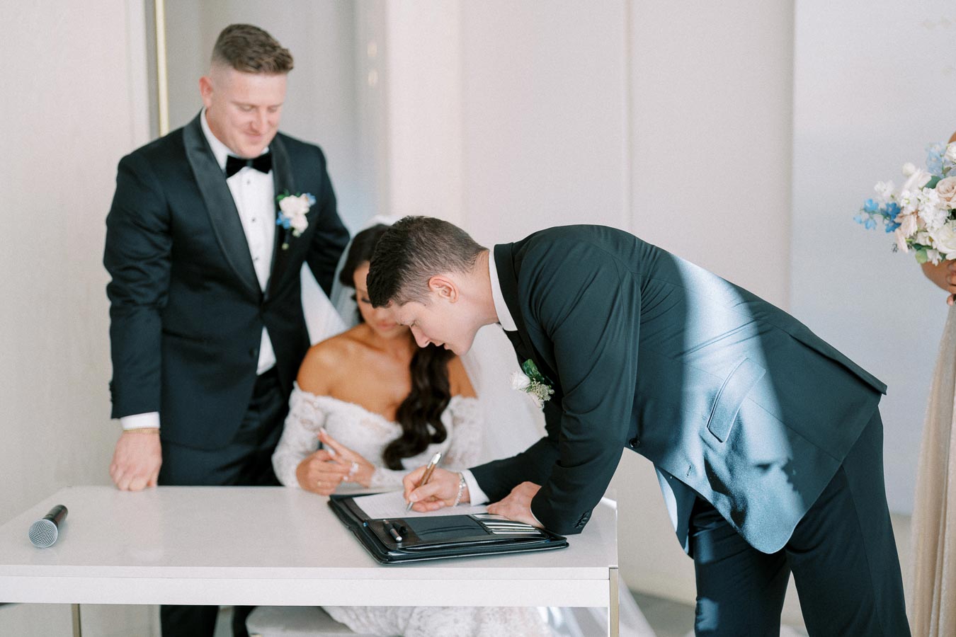 Groom signing wedding registry as bride and best man look on, dressed in elegant formal attire.
