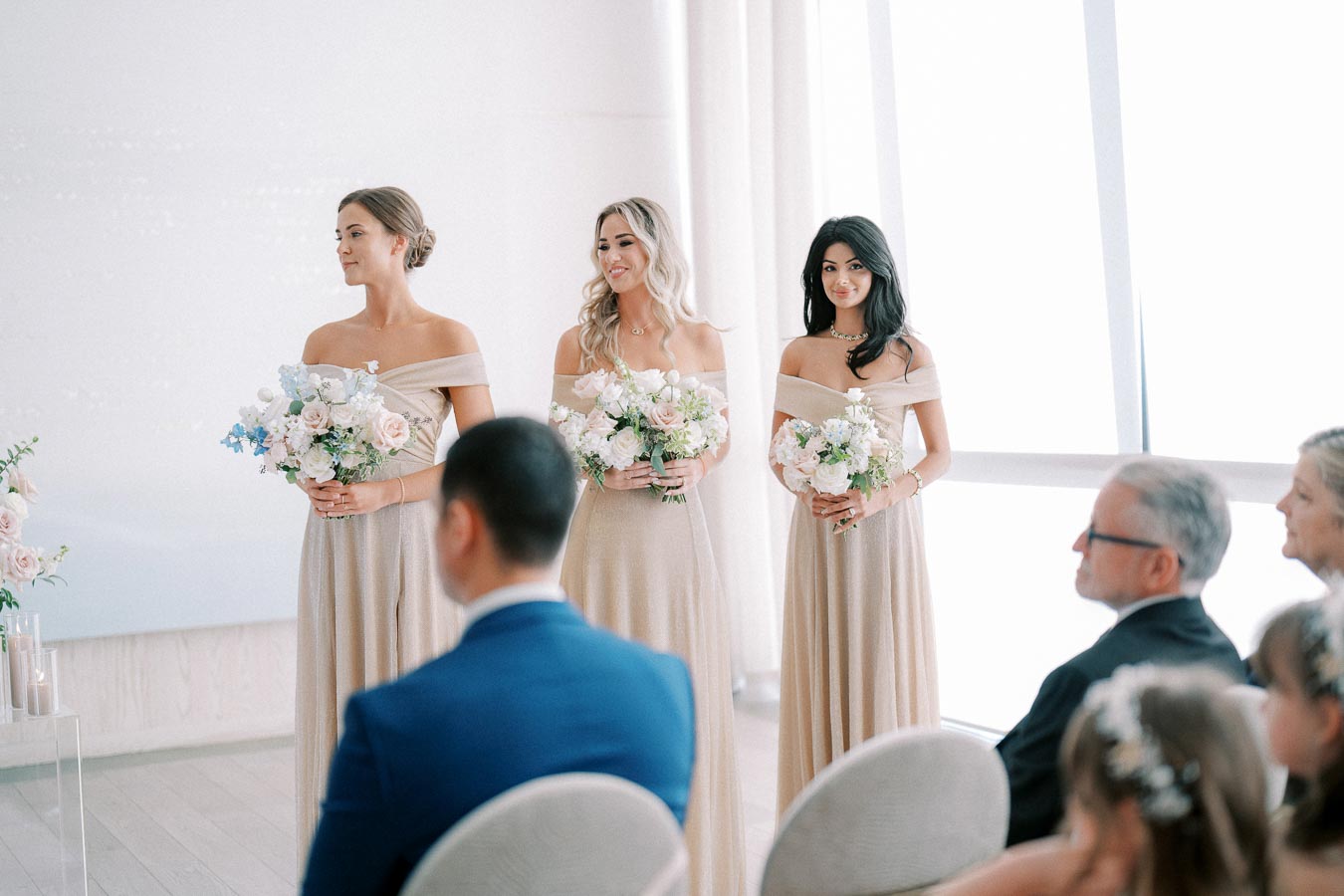 Three bridesmaids in elegant off-the-shoulder beige dresses holding floral bouquets stand in a bright, modern wedding venue, with seated guests in the foreground.