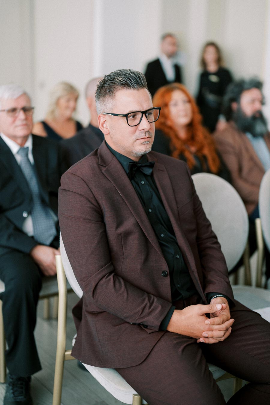 Man in a maroon suit with glasses sitting attentively during a formal event, surrounded by other well-dressed attendees.