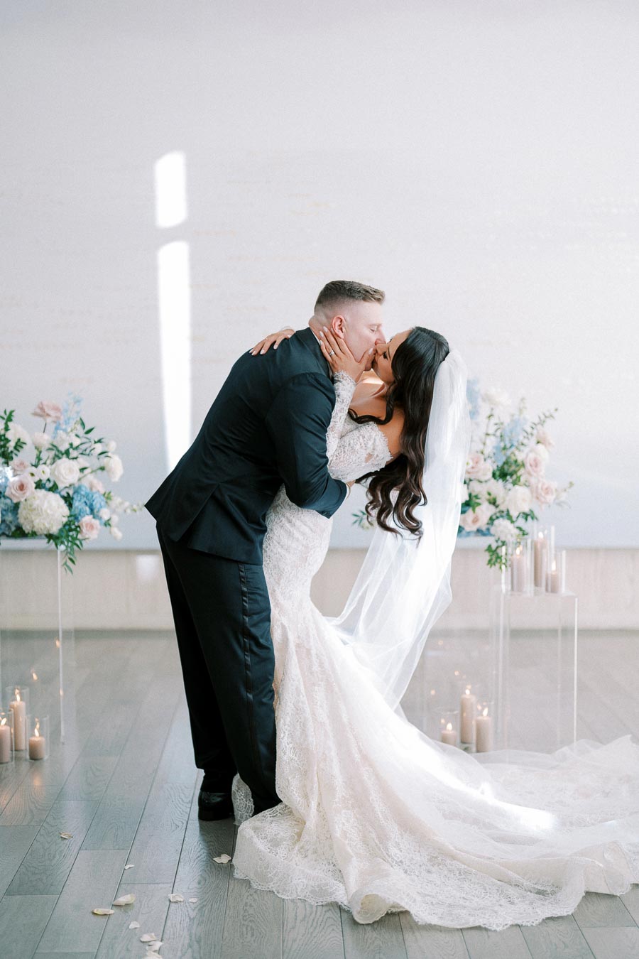 Elegant wedding couple sharing a romantic kiss, bride in lace gown and veil, groom in navy suit, surrounded by floral arrangements and candles.