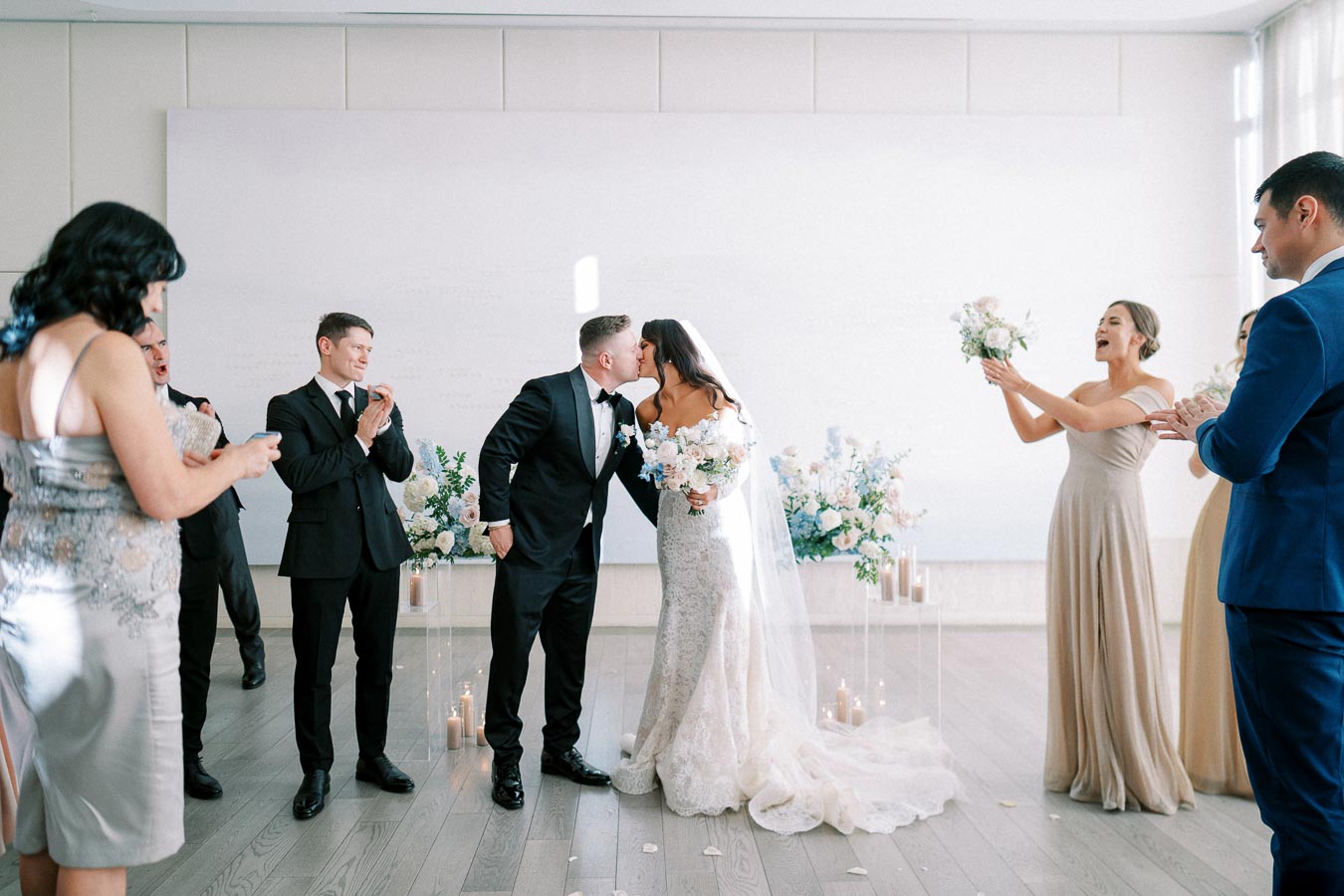 A newlywed couple shares a kiss during their wedding ceremony, surrounded by joyful bridesmaids and groomsmen. The bride is in an elegant lace gown holding a bouquet, while the groom wears a classic black tuxedo. The background features floral arrangements and candles, creating a romantic atmosphere.