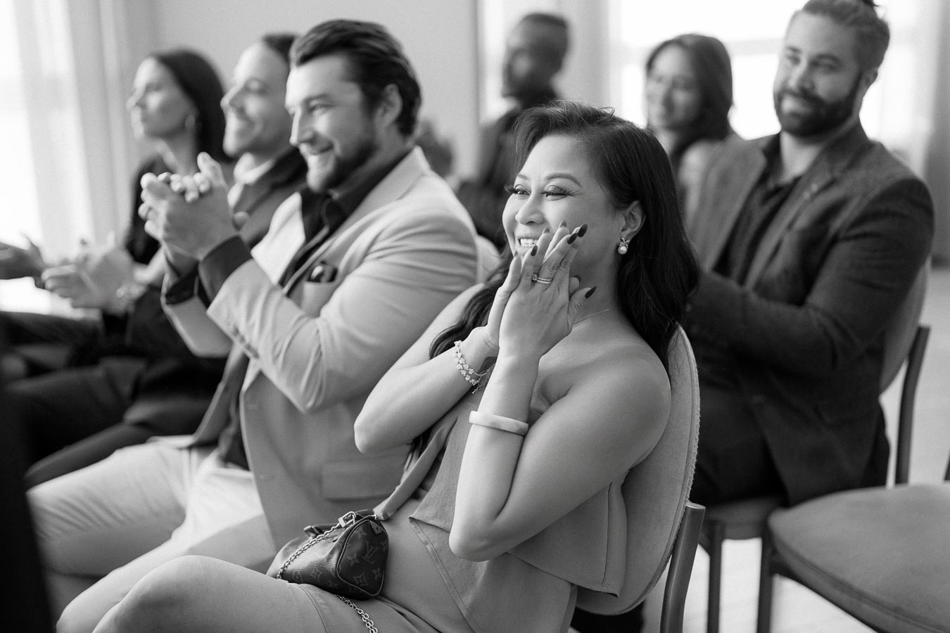A group of well-dressed people sitting and clapping enthusiastically at an event, with a focus on a smiling woman in a formal setting. Black and white photograph capturing audience engagement.