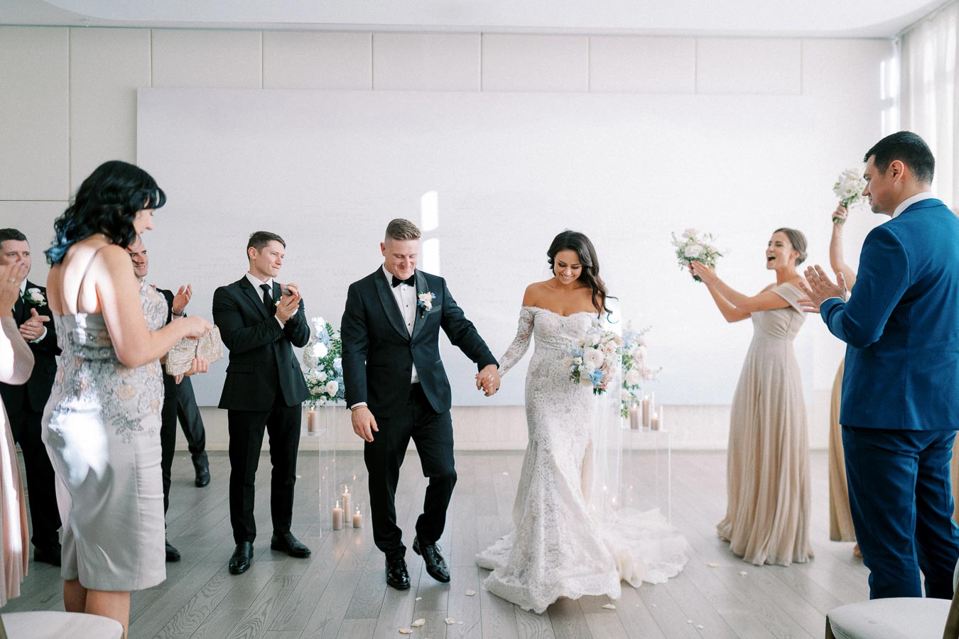 Newlywed couple joyfully exit their wedding ceremony, hand in hand, amidst applauding guests in an elegantly decorated venue. The bride wears a lace off-shoulder gown while the groom is in a black tuxedo, surrounded by floral arrangements and candles.
