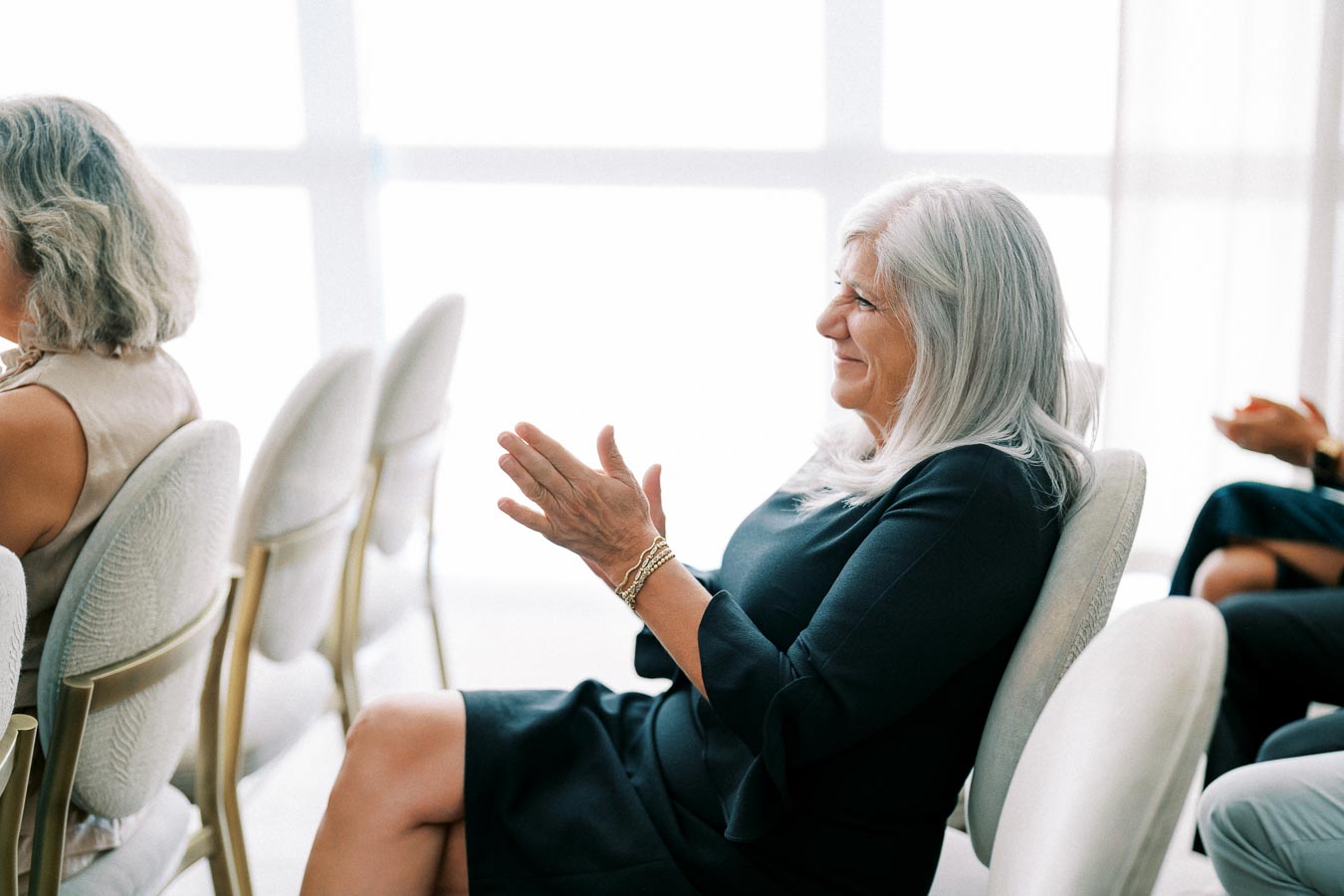Elderly woman with long gray hair smiling and clapping while sitting in an audience at an event.
