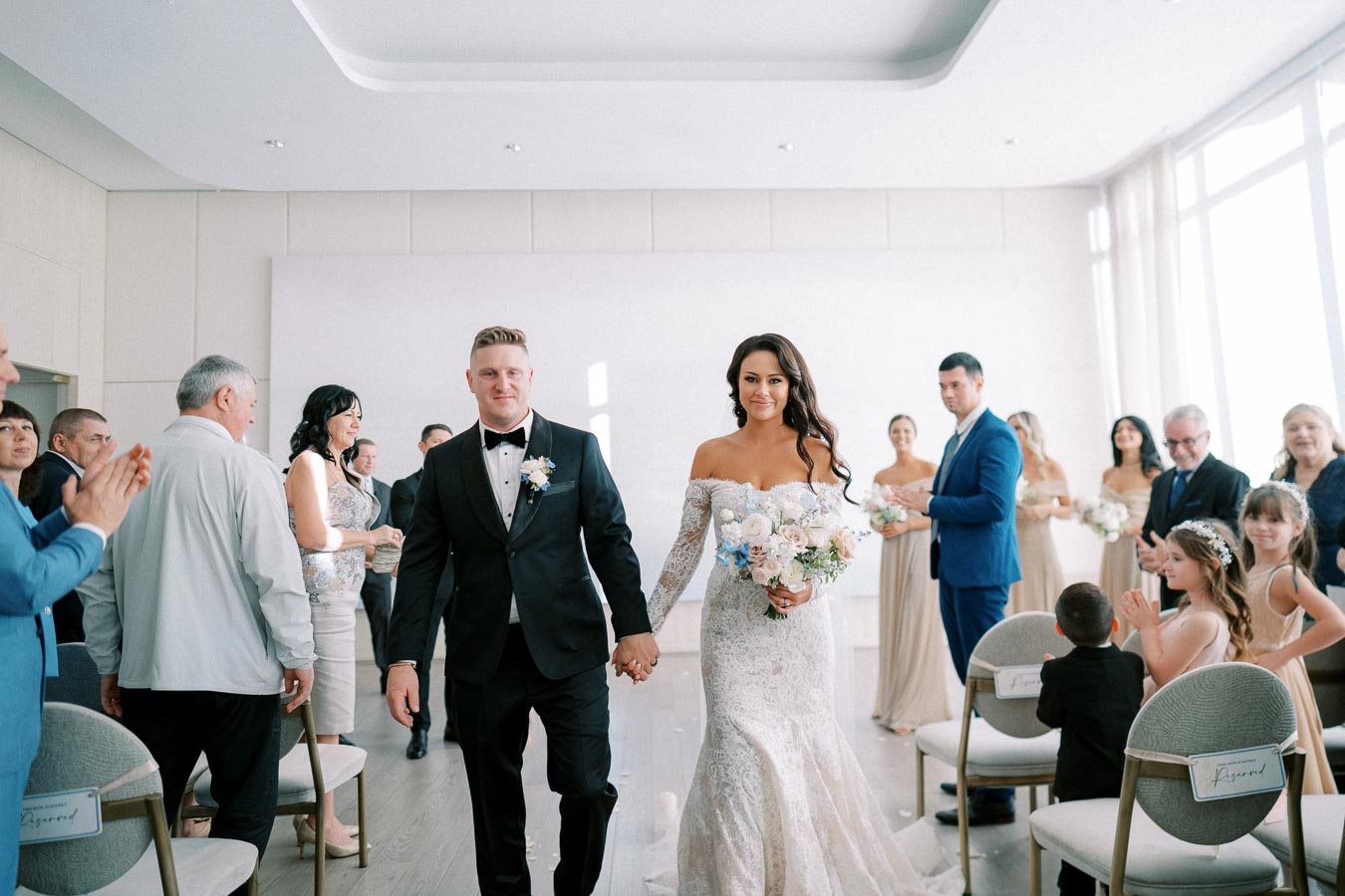 Bride and groom walking down the aisle during a wedding ceremony with guests celebrating and clapping in an elegantly decorated venue.