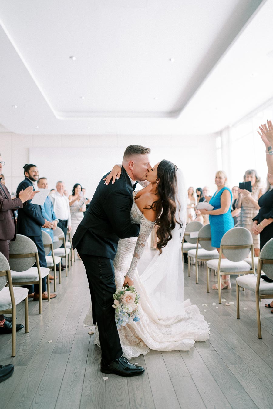 A newlywed couple shares a romantic kiss at their indoor wedding ceremony, surrounded by applauding guests. The bride, in an elegant lace gown holding a bouquet, is embraced by the groom in a black suit as they celebrate their marriage.