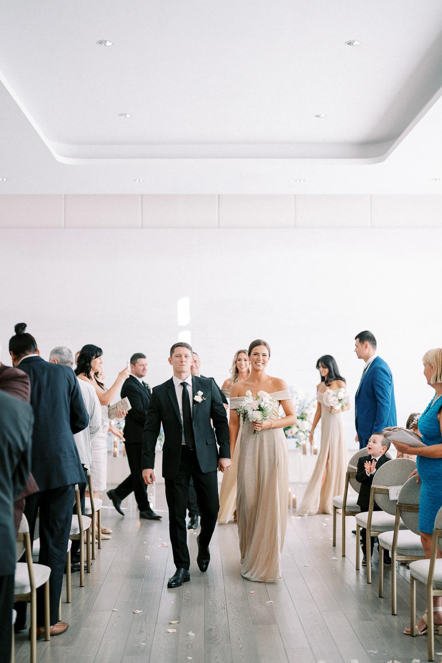 Alt text: Couple walking down the aisle at a wedding ceremony, surrounded by guests in formal attire, in a bright and elegant venue.