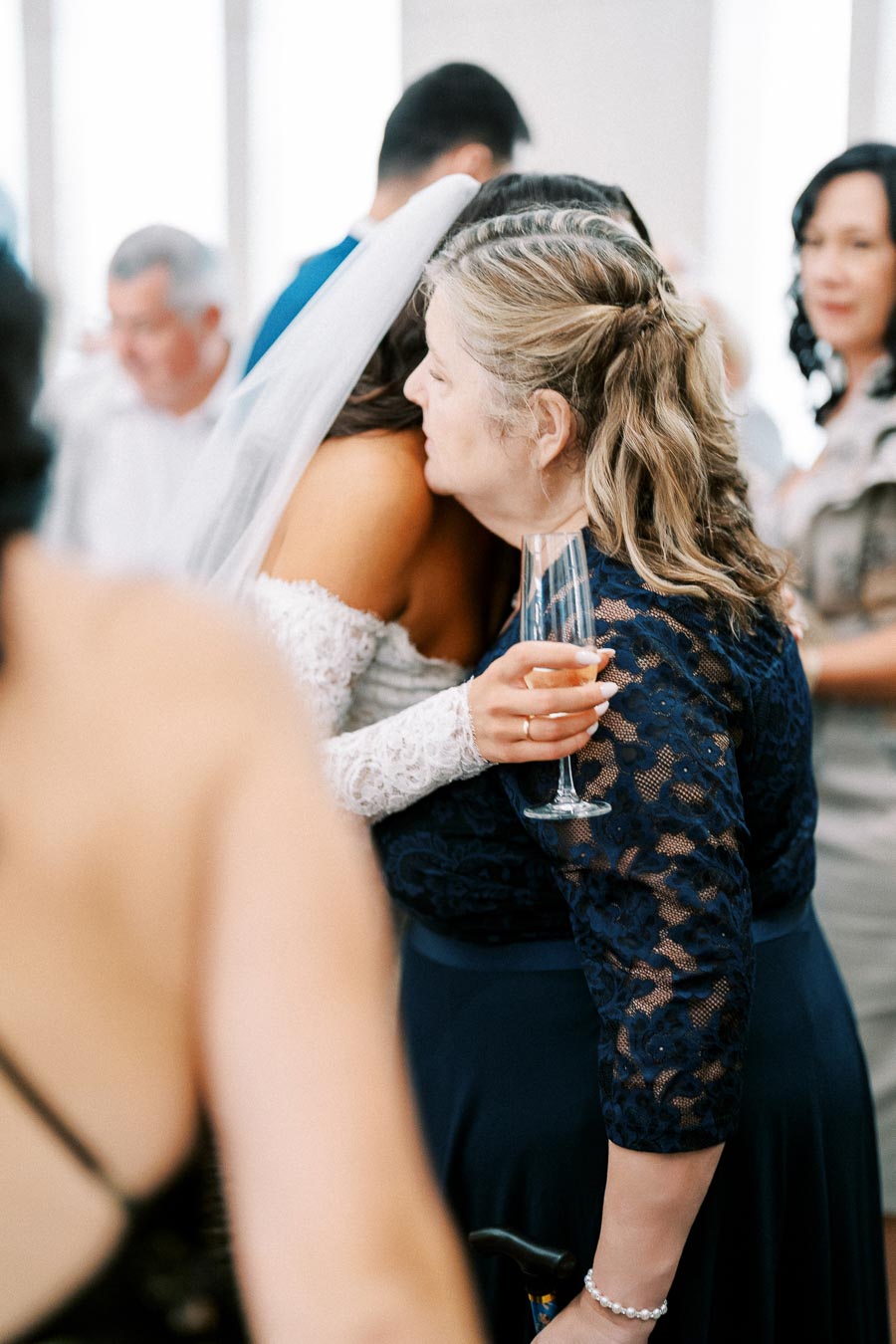 A bride in a white lace gown and veil hugs a woman in a navy lace dress holding a champagne flute at a wedding reception.