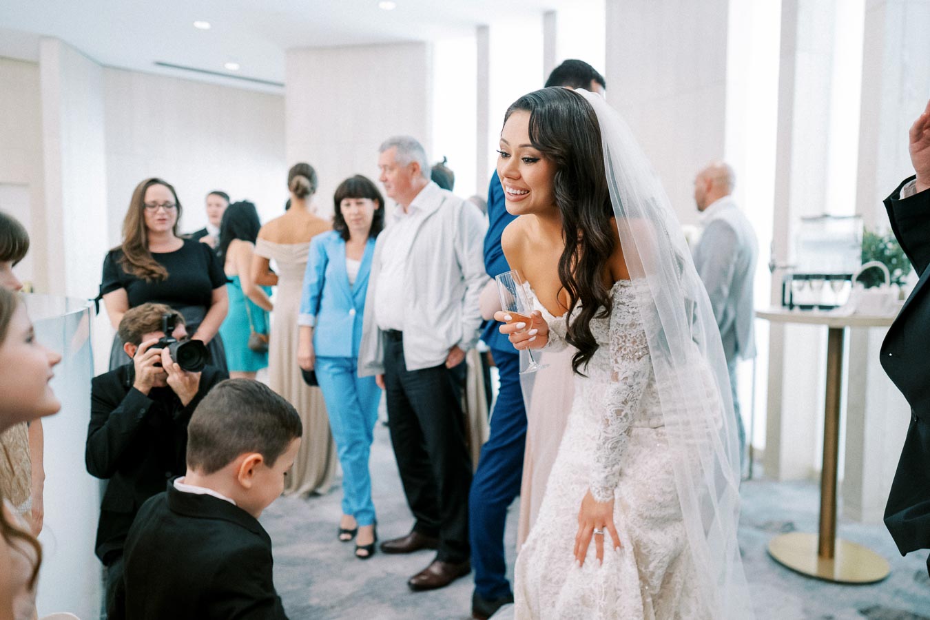 A bride in a white lace wedding dress smiles and interacts with guests during a reception, surrounded by people in formal attire and a photographer capturing the moment.