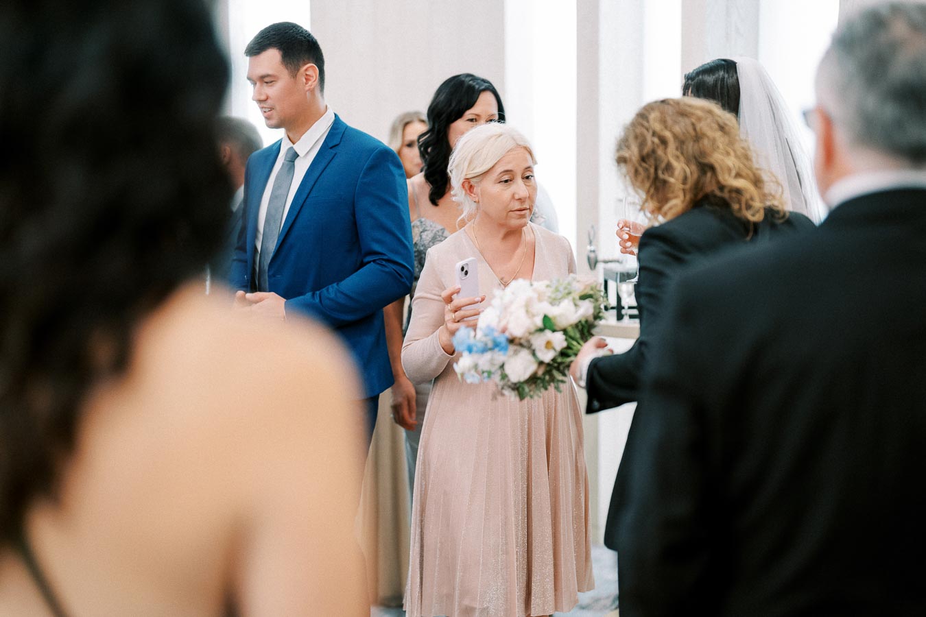 Guests mingling at an elegant indoor wedding reception, with a focus on a woman holding a bouquet and wine glass, surrounded by well-dressed attendees.