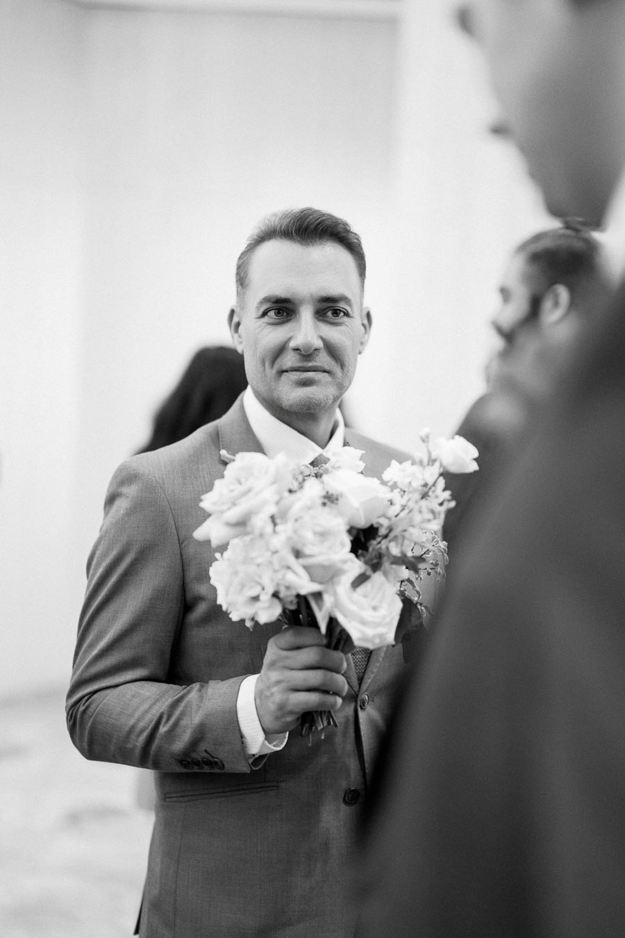 A groom in a suit holding a bouquet of flowers, captured in a black and white wedding setting.