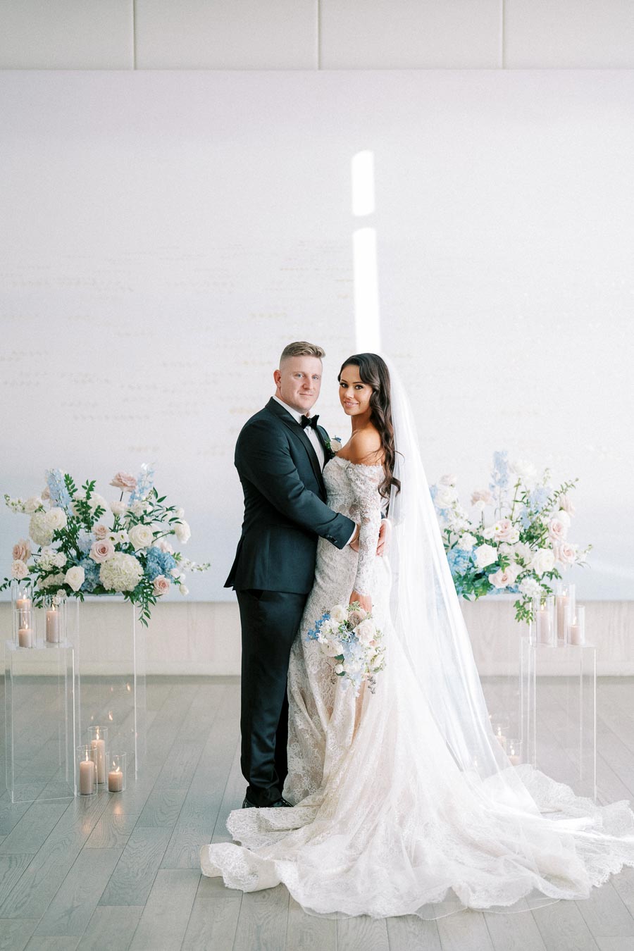 A bride and groom in elegant wedding attire pose together in a beautifully decorated venue with delicate floral arrangements and candles, symbolizing a romantic and sophisticated wedding ceremony.