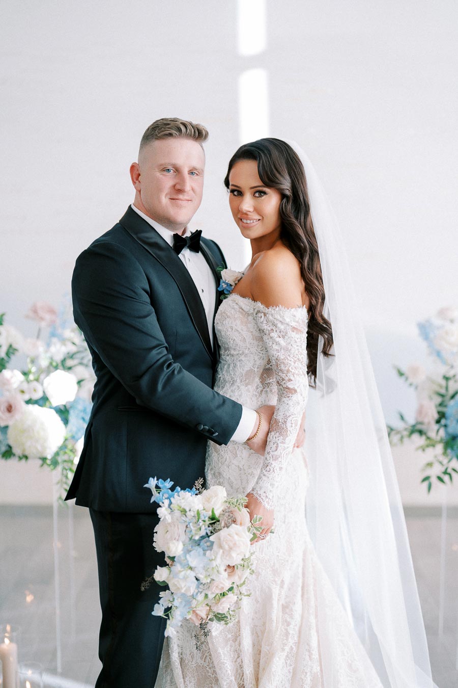 A bride and groom posing in an elegant setting, with the bride in a lace off-shoulder gown holding a pastel floral bouquet, and the groom in a classic black tuxedo.