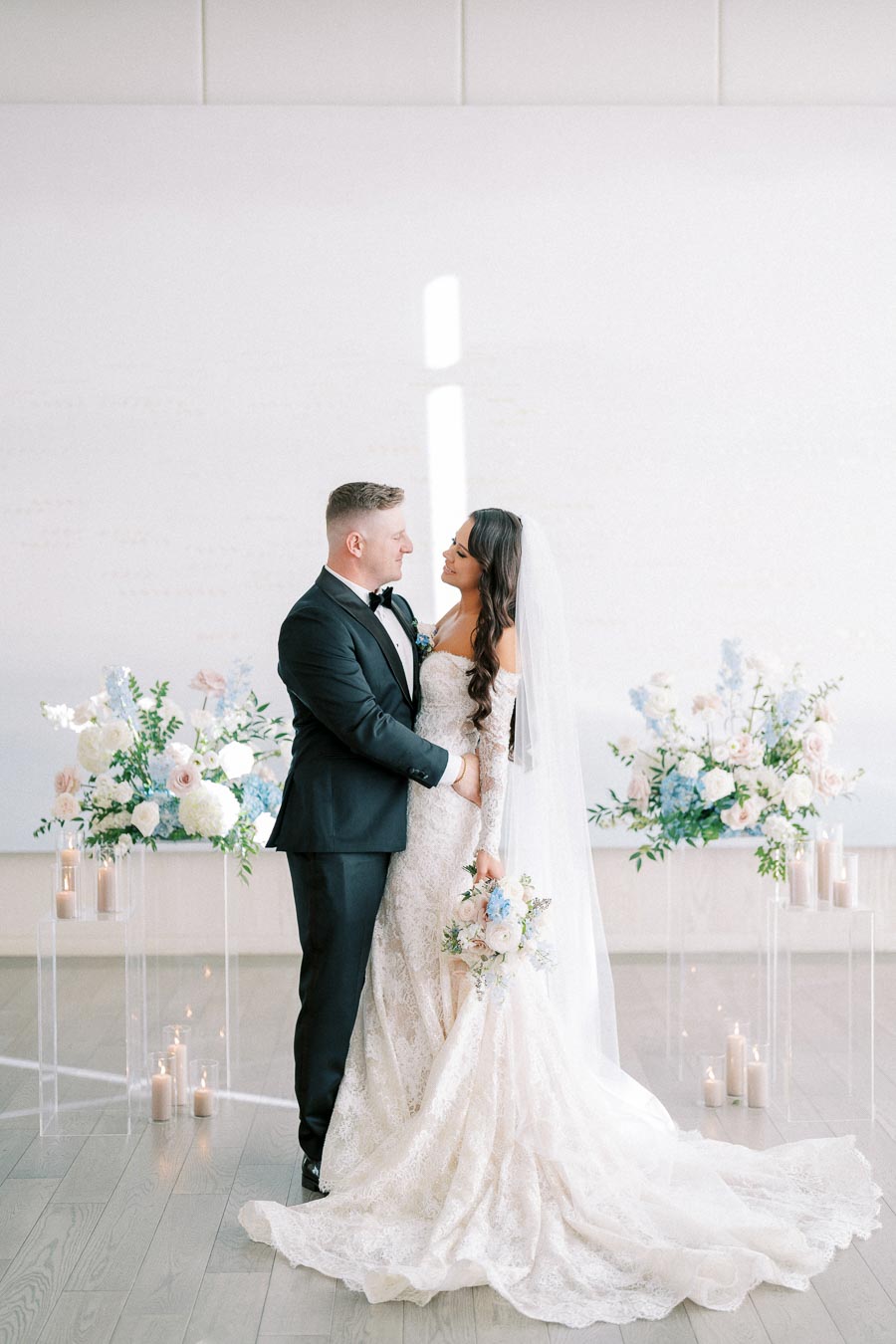 A bride and groom share a romantic moment during their wedding ceremony, surrounded by elegant floral arrangements and flickering candles, with the bride in a lace wedding dress and the groom in a classic black suit.