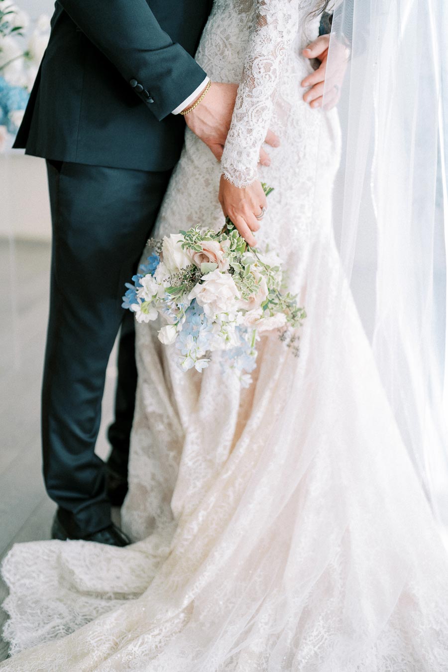 Bride and groom embrace in elegant wedding attire, with the bride holding a bouquet of white and blue flowers.