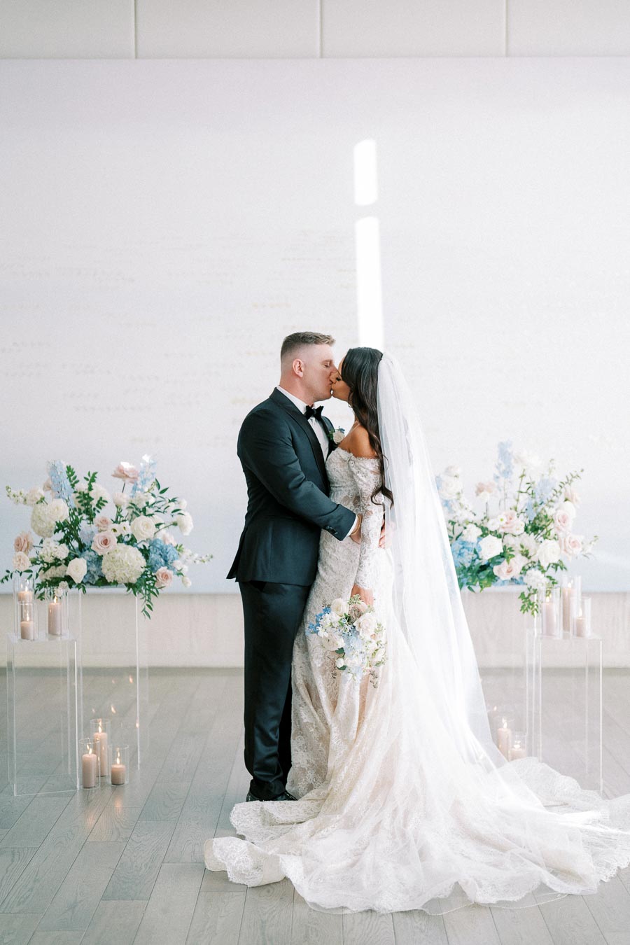 A bride and groom share a kiss during their elegant indoor wedding ceremony. The bride is in a lace gown and veil, holding a bouquet of pastel flowers. The groom is in a black suit. They are surrounded by floral arrangements and candles, creating a romantic atmosphere.