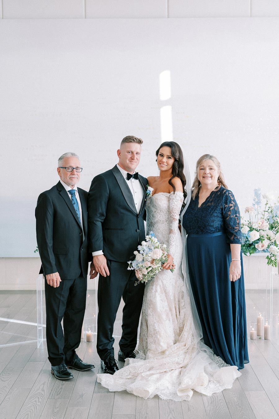 Elegant wedding portrait featuring a bride in a lace gown and groom in a tuxedo, posing with two older adults, likely parents, in formal attire. The group stands in a bright venue with floral arrangements and candles.